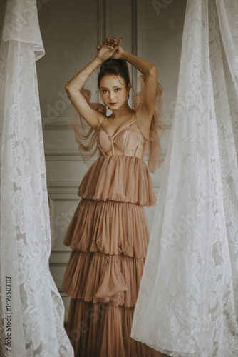Elegant woman in a blush-toned tiered gown poses between lace curtains indoors
