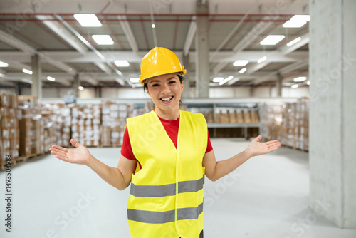 Female warehouse worker in yellow vest and helmet smiling with arms outstretched, standing in modern storage facility.