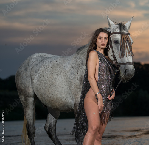 Beautiful brunette woman with long hair and brown scarf  rides  on a white horse in the river. Sensual and attractive girl  interacting and ride a horse in the water at sunset