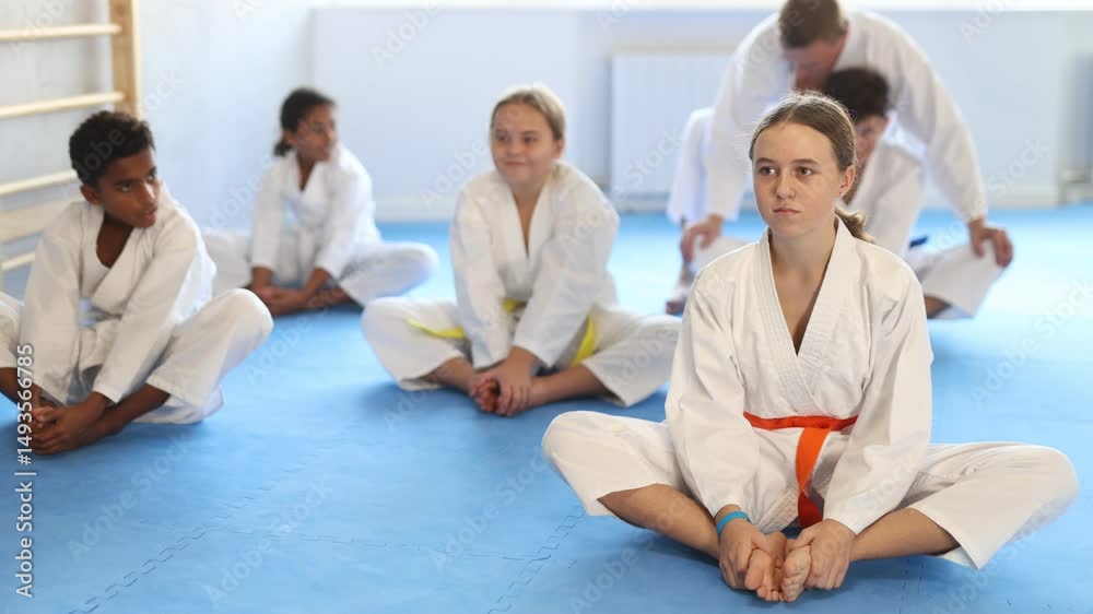 Group of positive tweens of different nationalities wearing white kimonos sitting on tatami in gym, performing butterfly stretch during martial arts workout with coach. High quality 4k footage