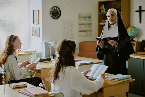 Elderly nun standing near desk holding book while instructing two female students. Books and study materials spread across desk in well-lit classroom setting
