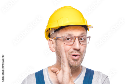 Middle-aged construction worker holding hand near mouth and telling secret, isolated on white background. Gossip concept. Craftsman against white wall