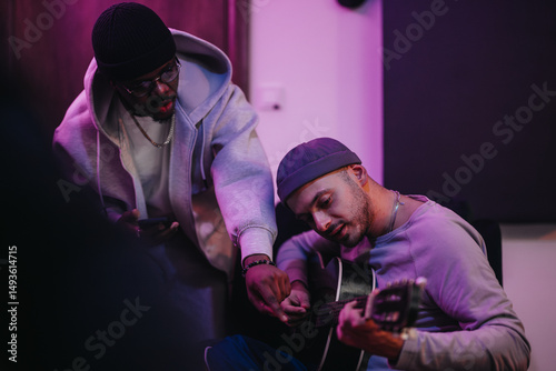 Musicians in a studio setting collaborate on music, highlighting the creative process. One plays the guitar while the other provides input, fostering a multicultural exchange in an artistic ambiance.