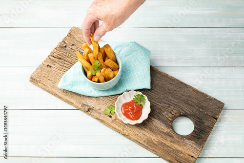 Hand taking a home-style French fry out of a blue bowl on a rustic cutting board.