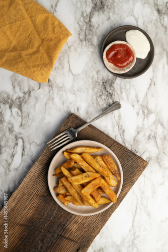 Crispy, golden, seasoned, homestyle french fries on a plate with a fork on wood cutting board and marbled background. Vertical
