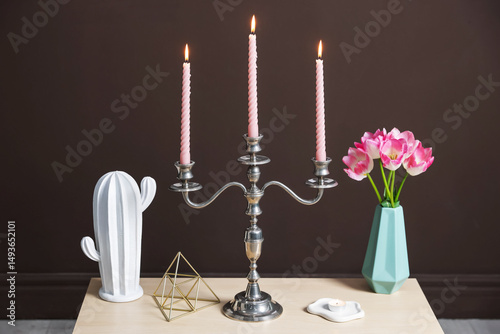 Elegant candlestick with burning candles and decor on table near brown wall