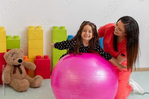Pediatrician assisting smiling girl balancing on pink exercise ball in clinic