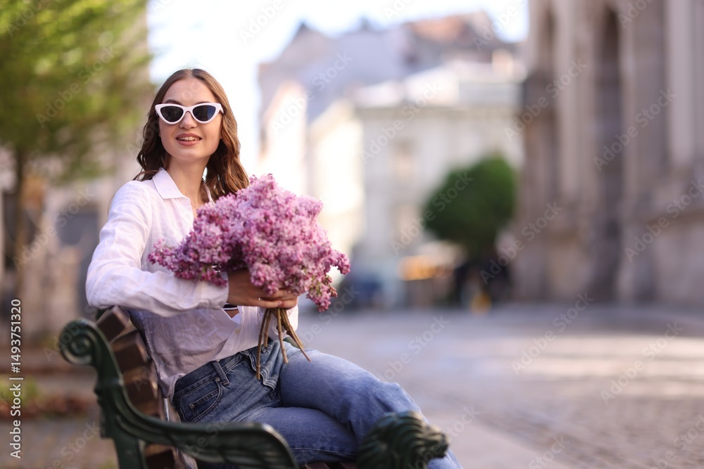 Fototapeta premium Smiling woman in sunglasses with bouquet of lilac flowers sitting on bench outdoors. Space for text