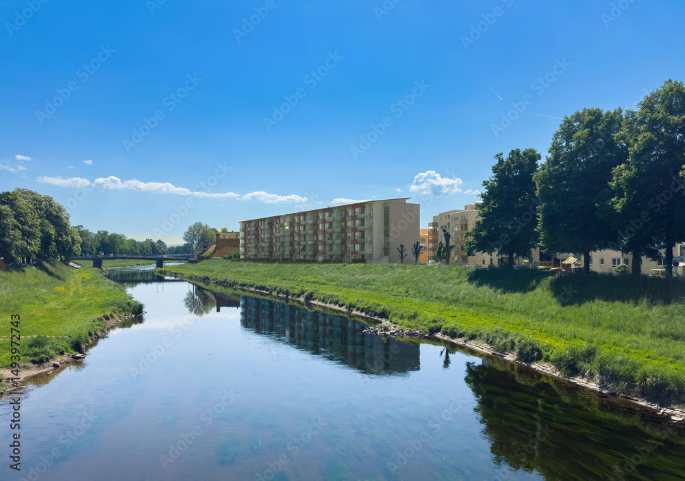 Fototapeta premium Scenic view of the Murg river in Rastatt, Germany with modern residential buildings on the right bank and lush greenery reflecting in the calm water under a bright blue sky