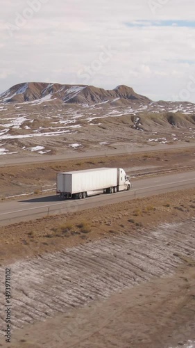 DRONE: White lorry hauling a heavy cargo container drives along a scenic interstate highway on sunny winter day. 18 wheeler drives towards the snowy landscape in the distance. Truck drives across Utah