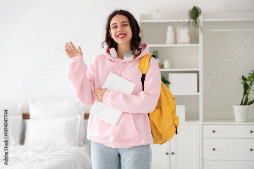 Female Indian student with backpack and laptop waving hand in bedroom