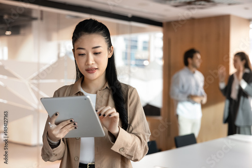 Focused Vietnamese businesswoman working on digital tablet device standing in meeting room, colleagues in background. Professional workflow using modern innovative business AI tool, apps and platforms