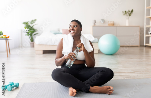 Obraz na plátně Plump black lady in sportswear sitting on sports mat with bottle of clear water at home, full length