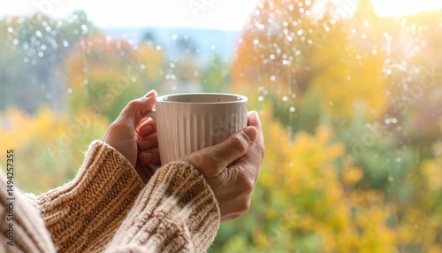 Cozy autumn scene with a person holding a warm beverage by a rain-soaked window overlooking colorful foliage