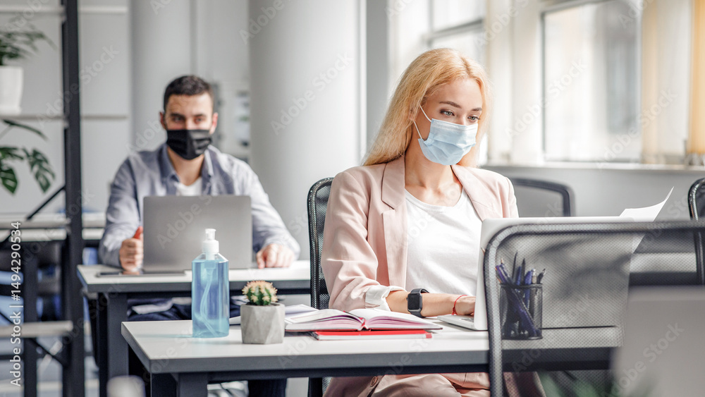 Fototapeta premium New working conditions and social distancing after quarantine. Blonde woman in protective mask works with documents and laptop at workplace with notepad and big bottle of antiseptic in interior