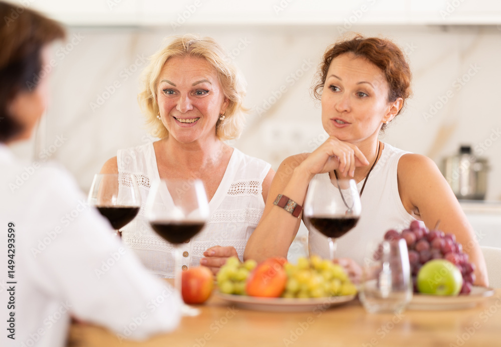 Relaxed adult female friends having fun at house party, sharing stories and laughing gathered around table with wine and appetizers in cozy kitchen Relaxed adult female friends having fun at house party, sharing stories and laughing gathered around table with wine and appetizers in cozy kitchen