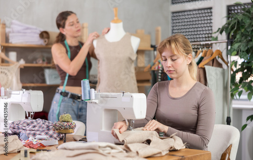 Skilful young tailoress using sewing-machine while other working with mannequin in sewing workshop