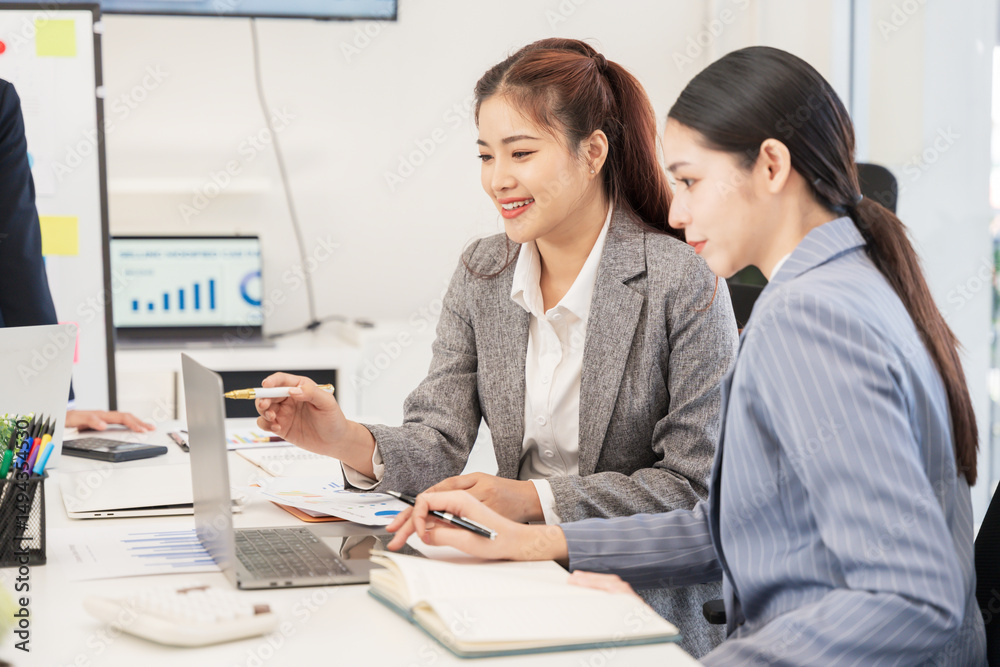 Two Asian businesswomen sit together working, using a laptop to check documents. Two Asian businesswomen sit together working, using a laptop to check documents.