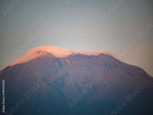 A majestic volcano is bathed in soft evening light. Wisps of clouds wrap its summit, glowing pink under the sunset sky, highlighting the rugged texture of its slopes