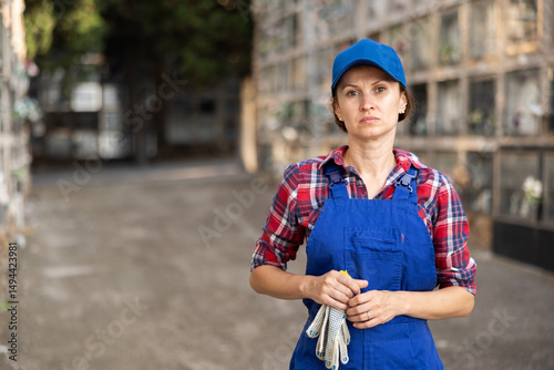 Wallpaper Mural Focused skilled female cemetery worker in blue overalls and cap standing near columbarium wall, holding gloves, during daily maintenance duties Torontodigital.ca