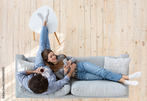 Young couple resting at home, using smartphone and drinking coffee while resting at home on couch, top view, free space