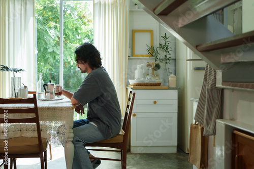 Fotografie Asian man sits at a dining table in a kitchen, holding a cup of coffee and enjoying a moment of solitude while sunlight streaming through the windows
