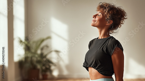 A woman in her early 30s, wearing casual activewear, standing in a soft morning light by a large window, stretching and smiling with eyes closed,