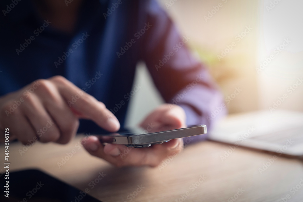 Fototapeta premium A man is using his cell phone to text. The phone is on a table next to a laptop