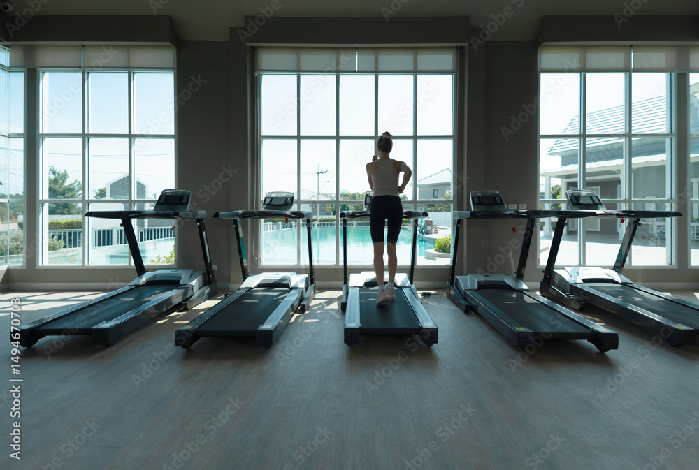 Fototapeta premium Young woman running on a treadmill in the gym. Stand facing large window, enjoy view of a pool outside. The bright sunlight fill the fitness area, creating an inviting atmosphere for exercise.