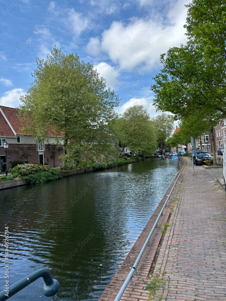 Fototapeta premium Modern Canal Living: Serene Waterway and Contemporary Dutch Homes A calm, wide canal reflects the bright sky, bordered by meticulously maintained green lawns and a clean