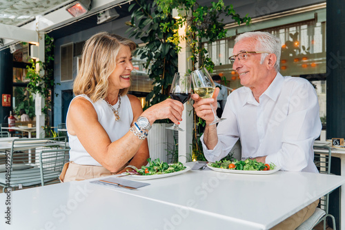 Mature retirees clinking wine glasses, sharing loving moment during outdoor dining, savoring gourmet meal while celebrating milestone together