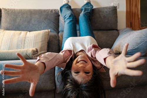 Young woman lying upside down on sofa is reaching out with open arms