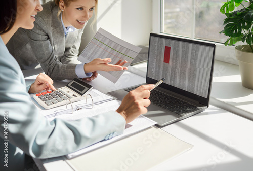 Two financial accountants analysing business spreadsheets on laptop computer, sitting at office desk, looking through paper documents, using calculator working with databases, checking excel tables photo