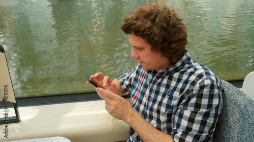 Man tourist looking at smartphone sitting in river tram public transport in city. Male reading news, surfing online, chatting with friends in mobile phone. Travel tourism trip journey voyage.