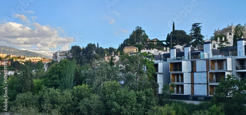 Modern apartment buildings with white and wood-paneled exteriors beside a green forest, set on a hillside with traditional homes in the background; apartment buildings.
