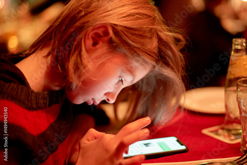 Child focused on smartphone, sitting at dinner table with drinks and plate nearby.