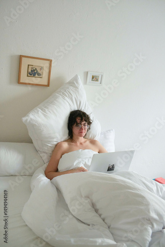 Young woman relaxing in white bedding while working on a laptop in a minimal bedroom.