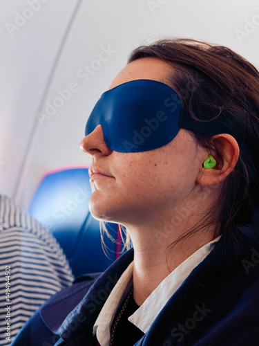A woman with an eye mask and earplugs sits on an airplane seat, appearing relaxed.