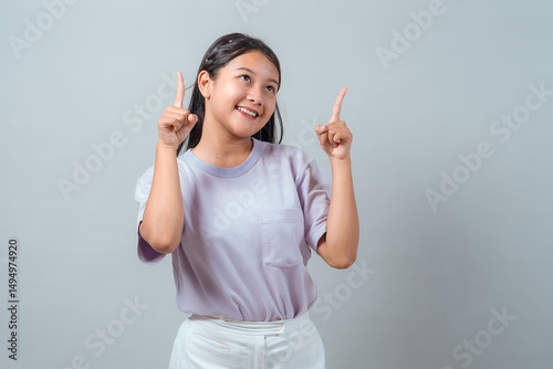 Cheerful young Asian girl in casual pastel outfit smiling and pointing upwards with both hands, posing against plain background, showing positive emotion.