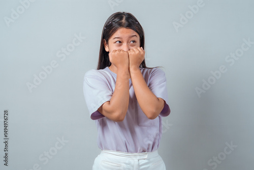 Shy young Asian woman expressing surprise or nervousness with both fists near her mouth, eyes looking sideways. Casual style, isolated on plain background, emotional expression.