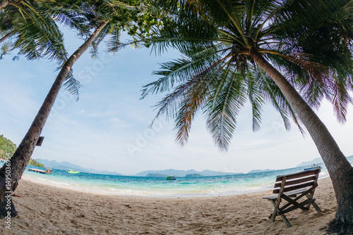 Fototapeta Naklejka Na Ścianę i Meble -  A beach scene with palm trees and a bench. The bench is empty. The sky is blue and the water is calm