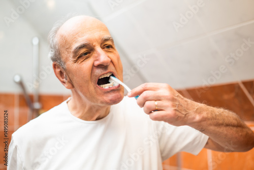 Portrait of senior man with gray hair brushing his teeth in front of the bathroom mirror.	