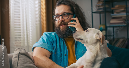 A man holds a phone to his ear and smiles as he speaks, all while his dog rests its head on his lap.