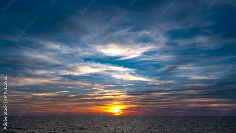 Fototapeta premium Dramatic sunset over the ocean. Vibrant colors paint the sky with clouds reflecting golden light on the water. Peaceful, serene scene