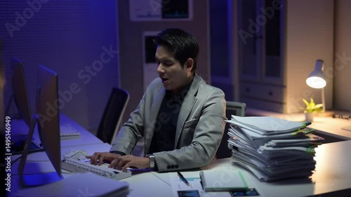 A man is sitting at a desk with a computer monitor and a stack of papers. He is typing on the keyboard and he is focused on his work. Scene is serious and focused