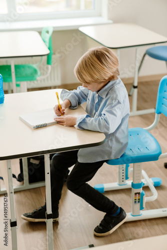 A schoolboy sits at a desk and writes with a pen in a notebook, at school, in the classroom