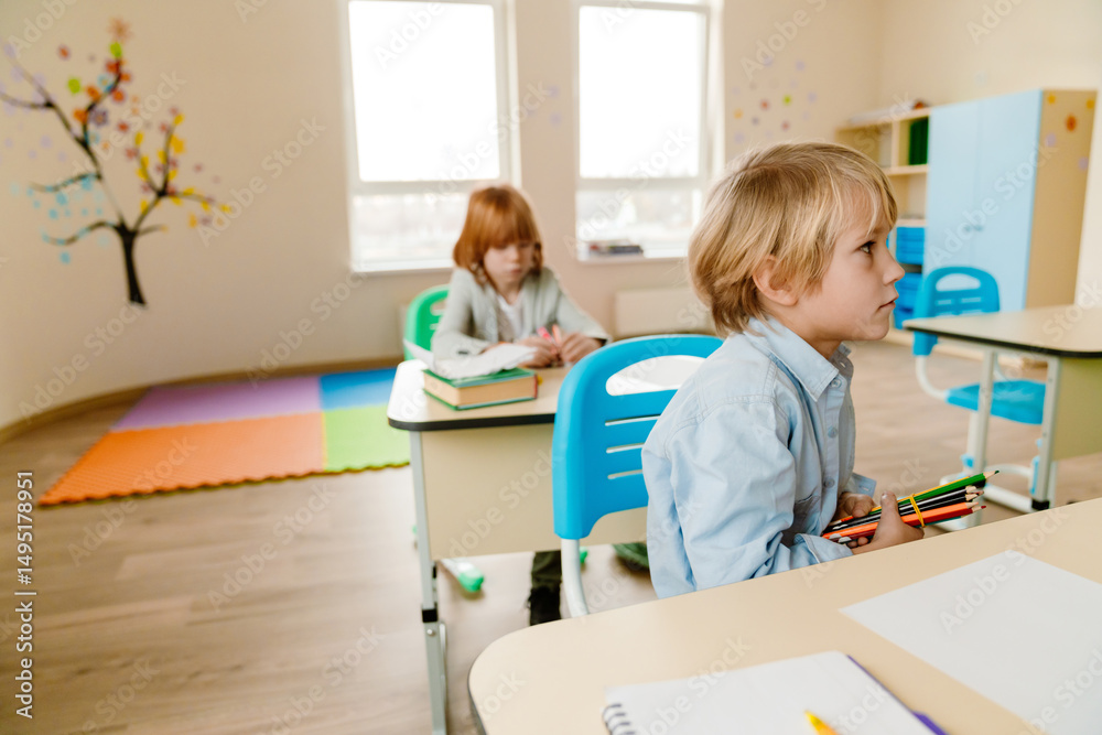 Obraz premium A schoolboy sits at a desk and looks in front of him while holding pencils, a classmate sits behind him at a desk, at school, in the classroom
