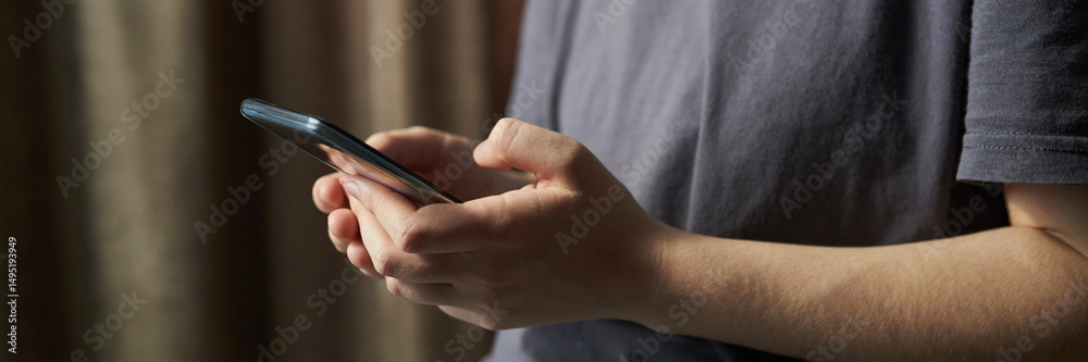Fototapeta premium Header of hands of person holding smartphone while standing indoors next to curtain