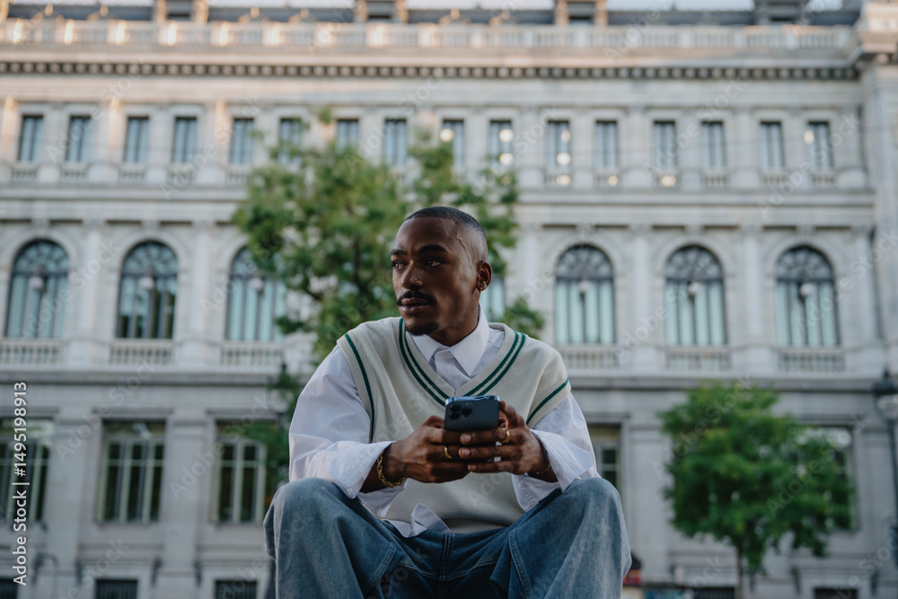 Obraz premium stylish Black male sits outside a historic building, holding his smartphone with a thoughtful expression.Young man modern outfit and confident posture reflect ambition and a digital lifestyle