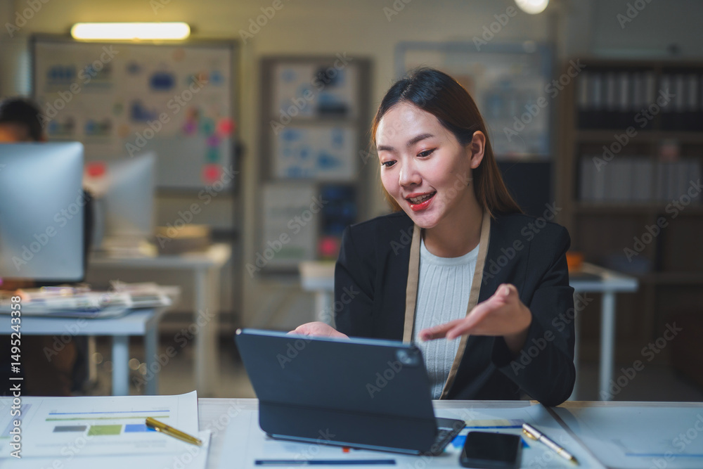 Young professional woman engaged in an online meeting, working late in a modern office, focused on her tablet and data analysis